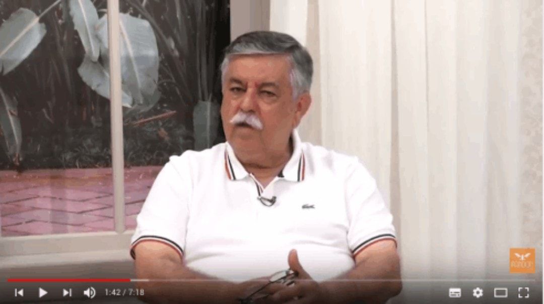 Um homem de bigode está sentado em frente a uma janela.